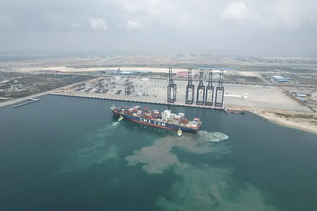 Ein Containerschiff navigiert im Lagos Port, assistiert von Schleppern. Kräne und Container deuten auf geschäftige Logistik hin.