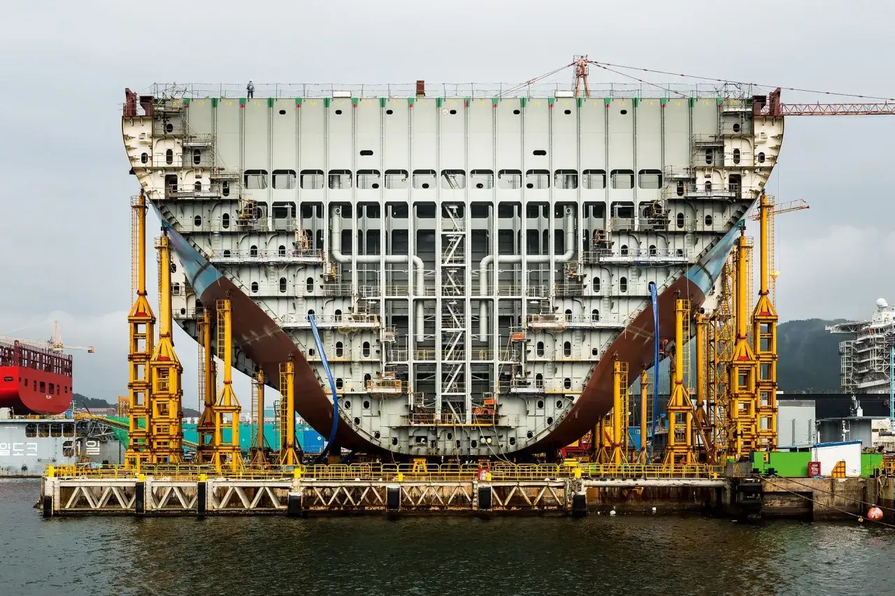 Gigantischer Schiffsrumpf im Bau, gestützt von gelben Gerüsten. Ein Arbeiter auf dem Deck, der die stowage (Lagerung/Stauung) im Vergleich zur storage (Lagerung/Aufbewahrung) im Schiffbau veranschaulicht.