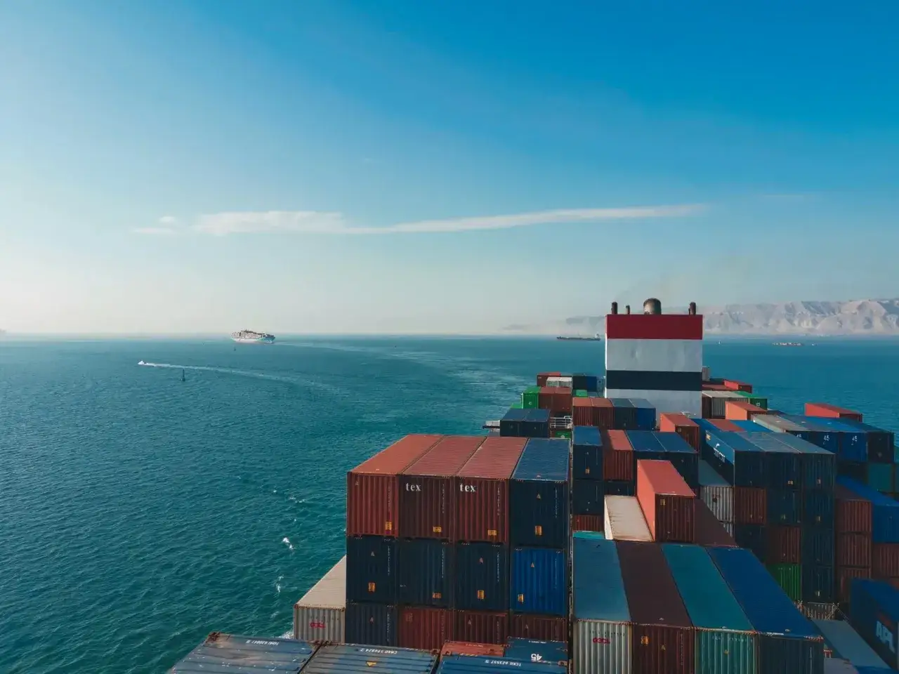 Ein riesiges Containerschiff, die CMA CGM Barracuda, f&auml;hrt auf dem Meer. Berge im Hintergrund, ein kleineres Schiff im Vordergrund.