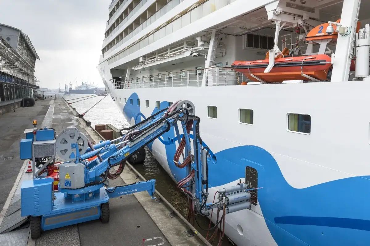 Ein Schiffsausrüster bereitet ein Kreuzfahrtschiff vor. Ein blauer Roboterarm reinigt die Bordwand.