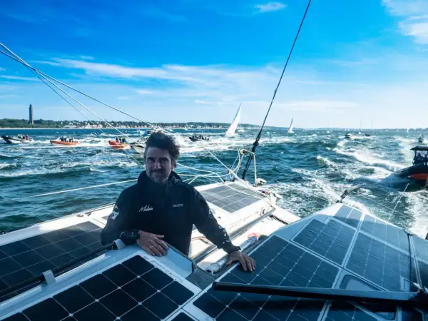 Boris Herrmann in Kiel, auf seinem Boot mit Solarpaneelen, umgeben von anderen Booten und Segelschiffen auf dem Meer.