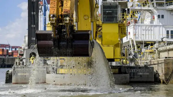 Gelbe Baggerschiff-Arme entleeren Wasser und Sedimente ins Meer. Ein riesiger Eimer schaufelt Material aus dem Wasser.