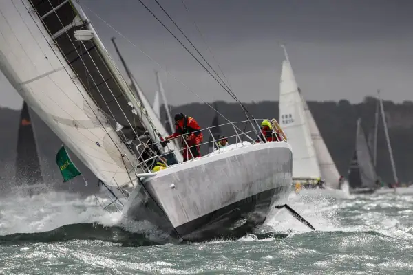 Segelboote kämpfen sich durch raue Wellen beim Fastnet Race. Ein Segler in Rot hält sich fest, während die Gischt aufspritzt.