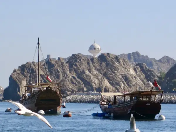 Traditionelle Dhaus-Boote im Oman Port, mit felsigen Bergen und einem weißen Kuppelgebäude im Hintergrund.