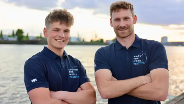 Melwin Fink und sein Teamkollege posieren lächelnd am Wasser. Sie tragen marineblaue Poloshirts mit dem Aufdruck "Around The World - Next Generation".