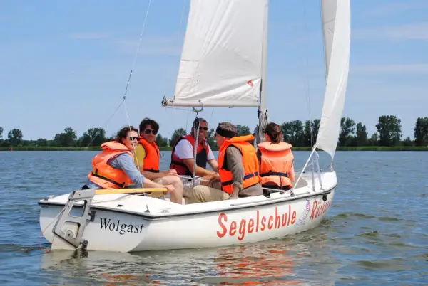 Fünf Personen in orangefarbenen Schwimmwesten genießen das Jolle segeln auf einem sonnigen See.
