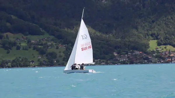 Segel reffen auf einem Boot mit drei Personen. Im Hintergrund bewaldete Hügel mit Häusern.