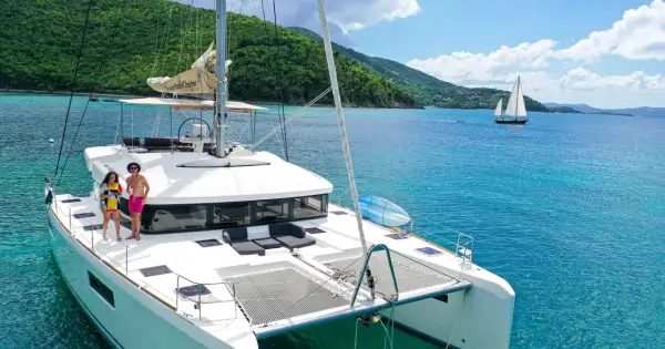 Ein Katamaran mit zwei Rümpfen, zwei Personen auf dem Deck, türkisfarbenes Wasser, grüne Hügel im Hintergrund.