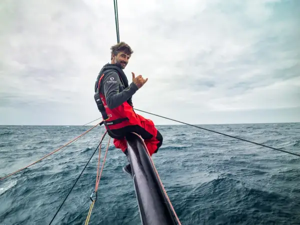 Boris Herrmann winkt lässig vom Bug seines Bootes, umgeben von rauer See und Segeltrossen.