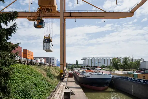 Ein Kran hebt einen Container im Hafen der Schweiz. Ein Frachtschiff liegt am Kai, umgeben von weiteren Containern und Lagerhallen.