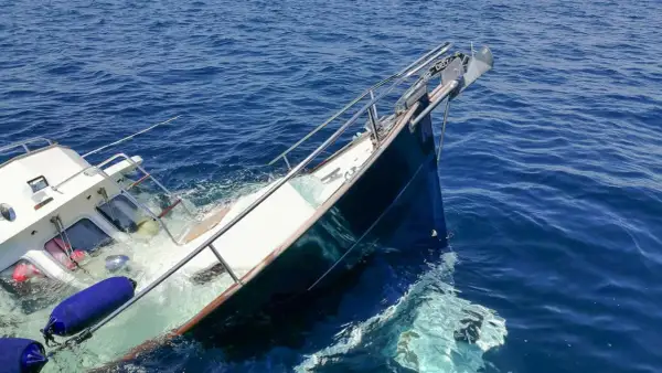 Ein sinkendes Boot neigt sich gefährlich im tiefblauen Meer. Rettungsringe und Fender treiben im Wasser.