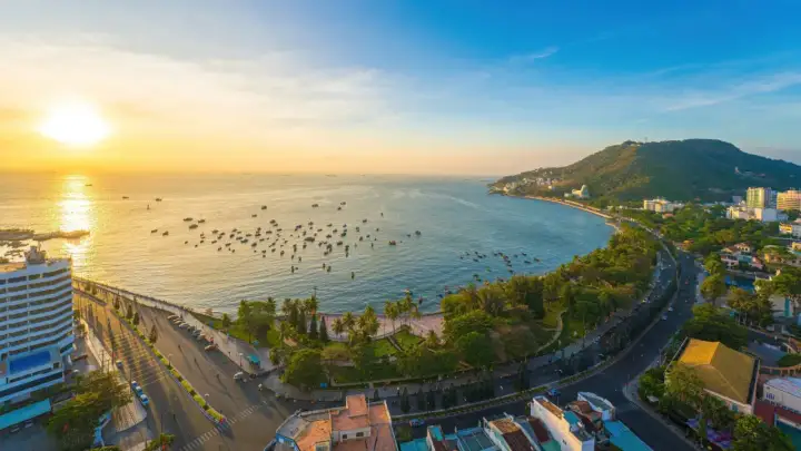 Sonnenuntergang über dem Vung Tau Port mit vielen Booten im Wasser und einer grünen Hügelkette im Hintergrund.